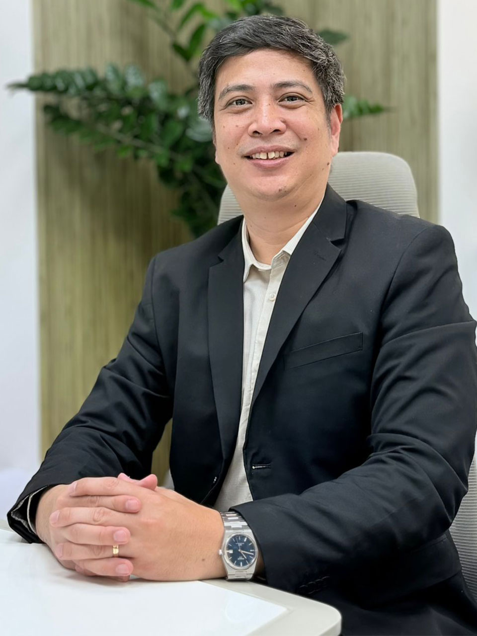 Professional portrait of a man in a black blazer sitting at a desk, hands folded, smiling at the camera with a watch visible on his left wrist.